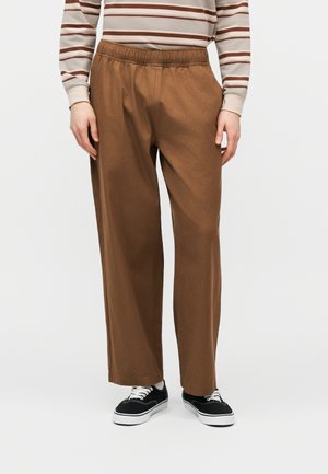 Personne portant un pantalon marron à jambes larges avec une ceinture élastique, une chemise à rayures à manches longues et des baskets basses noires avec des semelles blanches.