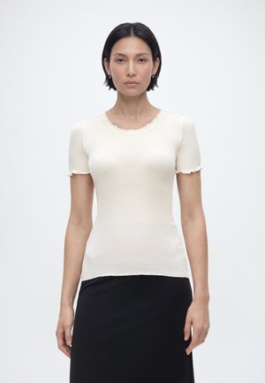 Rövid sötét hajú nő, aki testhezálló krémszínű rövid ujjú felsőt és fekete szoknyát visel, egyszínű fehér háttér előtt áll.