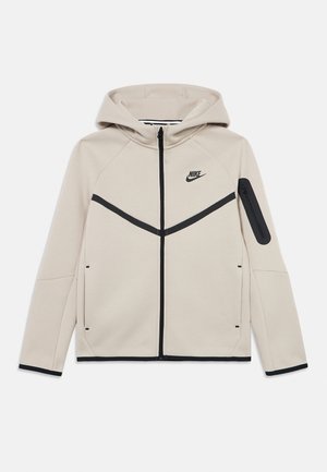 Jasnobeżowa bluza Nike z kapturem, z przednim zamkiem błyskawicznym, czarnymi akcentami, zakrzywionymi liniami wzoru oraz kieszenią na zamek na lewym rękawie. Miękka w dotyku.