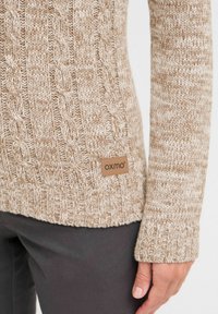 Beige gestrickter Pullover mit Zopfmuster, mit einem gerippten Saum und Bündchen. Das Markenlabel ist an der unteren Seite sichtbar.