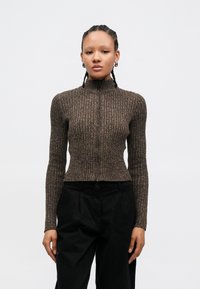 Brązowy sweter prążkowany z wysokim kołnierzem i zamkiem na przodzie. Posiada długie rękawy i dopasowany krój, w połączeniu z czarnymi spodniami.