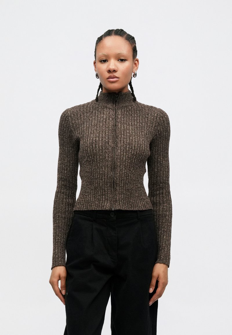 Brauner, gerippter Pullover mit hohem Kragen und vorderlichem Reißverschluss. Verfügt über lange Ärmel und eine figurbetonte Silhouette, kombiniert mit schwarzen Hosen.