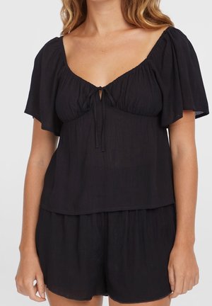 Mujer con una blusa negra de manga corta y holgada con un lazo delantero y pantalones cortos negros a juego, mostrada desde el cuello hasta la mitad del muslo.