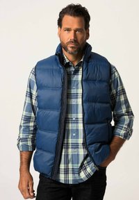 Sininen pehmeä liivi vetoketjulla, jossa on korkea kaulus ja sivutaskut, päällä vihreä ja sininen ruudullinen paita. Tummat farkut näkyvillä.