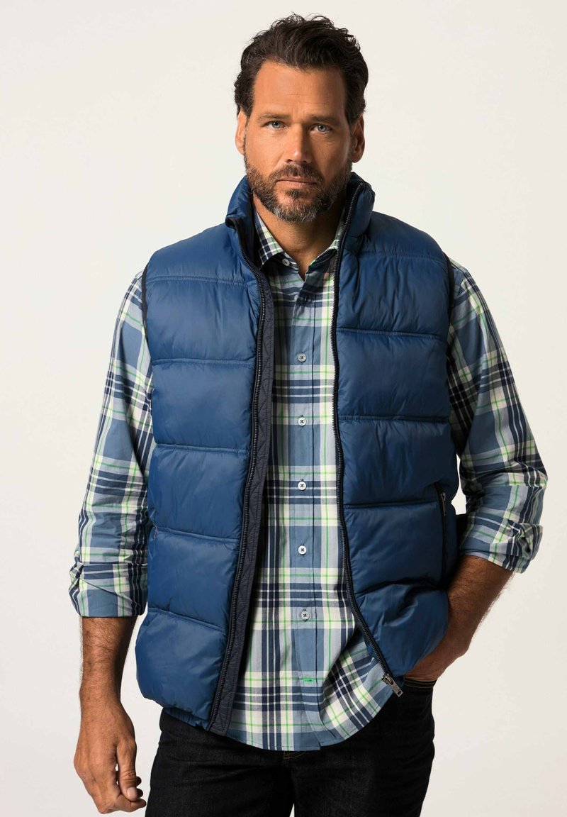 Sininen pehmeä liivi vetoketjulla, jossa on korkea kaulus ja sivutaskut, päällä vihreä ja sininen ruudullinen paita. Tummat farkut näkyvillä.