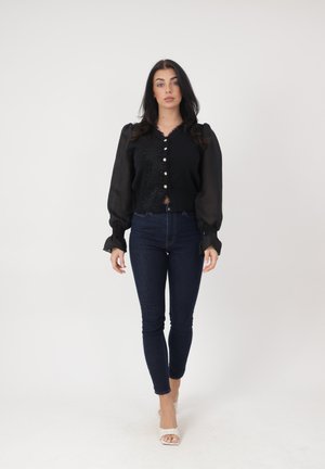 Frau trägt eine schwarze Bluse mit langen Ärmeln und Knöpfen, dunkle enge Jeans und weiße offene High Heels, steht vor einem schlichten weißen Hintergrund.