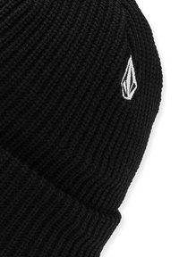 Bonnet en tricot noir avec une texture côtelée, présentant un petit accent logo blanc sur le côté. Le matériau semble doux et chaud.