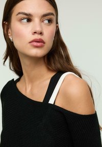 Pull noir à épaules dénudées avec tricot texturé, comportant des bretelles blanches et noires. Le design met en valeur les épaules nues et offre une coupe décontractée.