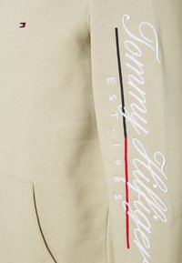 Sudadera con capucha beige con bolsillo canguro, texto bordado "Tommy Hilfiger" y líneas en contraste negras y rojas a lo largo de la manga.