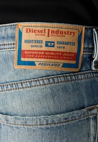Calças de ganga azuis com um rótulo de couro retangular. O rótulo apresenta texto vermelho e azul: "Diesel Industry," "Jeans de Qualidade Superior," e "Regulado."