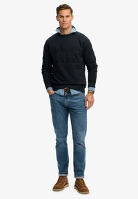 Sort sweatshirt med hævet tekst, lyseblå denimskjorte under, blå jeans og brune snørestøvler. Enkel, afslappet outfitdesign.