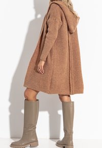 Gestrickter oversized Cardigan in Hellbraun mit Kapuze, strukturiertem Muster, überschnittenen Schultern und seitlichen Schlitzen. Kombiniert mit grauen knielangen Stiefeln.