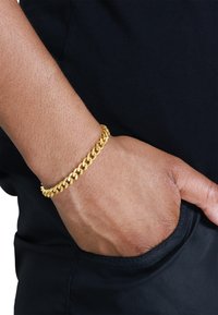 Goldene Kettenarmband am Handgelenk, mit einem klobigen Design und ineinandergreifenden Gliedern. Glatte Textur, reflektierende Oberfläche vor einem dunklen Hintergrund.