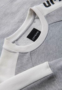 Sudadera gris con una mezcla de texturas, detalles en gris claro y blanco, puños acanalados y una marca "ombre" en negro en el hombro.