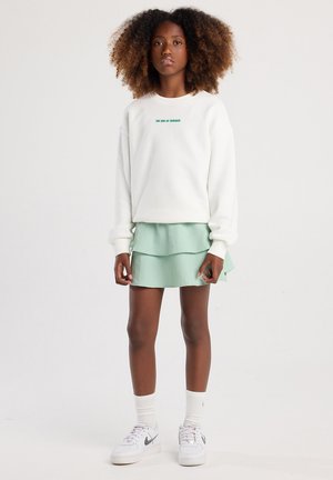 Sudadera blanca con texto verde, falda mint verde en capas con tejido texturizado, zapatillas blancas con acentos grises y calcetines blancos acanalados.