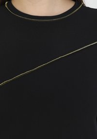 Schwarzes Oberteil mit rundem Halsausschnitt und diagonaler goldener Nahtdetail über der Brust, aus einem glatten, weichen Stoff gefertigt.