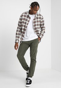 Rutete skjorte i beige, rød og marineblå; hvit T-skjorte; olivengrønne bukser; svarte sneakers med hvite såler og snøring.