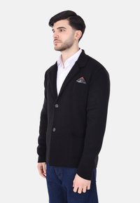 Cardigan nero a maglia con due bottoni, colletto a risvolto e tasca frontale con un fazzoletto da taschino decorato. Indossato su una camicia bianca.