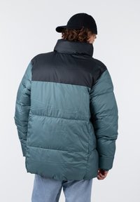 Veste matelassée en teal avec une section supérieure noire. Présente un col haut, un matelassage en canaux et une texture lisse. Manchons réglables.