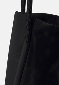 Svart tote-väska med en texturerad yta, med tjocka svarta flätade handtag och en öppen topp. Materialet verkar lätt och matt.