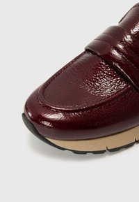 Chaussure en cuir bordeaux avec finition brillante, présentant un bout incurvé, une surface texturée et une semelle en caoutchouc beige contrastante. Détail de bride ajustable.