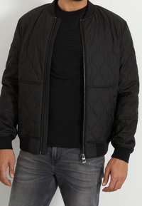 Chaqueta bomber negra acolchada con puños y dobladillo acanalados, con cremallera frontal y bolsillos laterales, llevada sobre una camiseta negra y jeans grises.