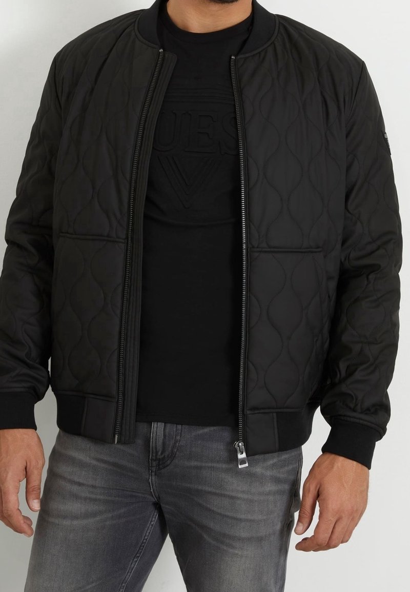 Chaqueta bomber negra acolchada con puños y dobladillo acanalados, con cremallera frontal y bolsillos laterales, llevada sobre una camiseta negra y jeans grises.