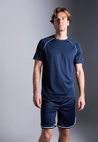 Jovem em pé contra um fundo cinza, vestindo uma camiseta atlética azul-marinho e calções combinando com detalhes brancos.