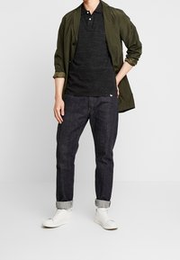 Veste légère olive, polo noir, jeans foncés en denim et baskets blanches. Coupe décontractée avec des manches retroussées et des textures discrètes.