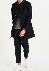 Manteau long noir, léger, au design simple, doté de boutons et d'un logo de marque visible. Porté sur un pull gris, associé à un pantalon noir et des baskets blanches.