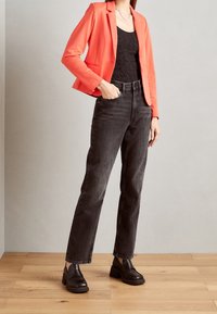 Blazer corail, top en dentelle noire, jean noir taille haute, et mocassins noirs épais. Sol en bois naturel en arrière-plan.