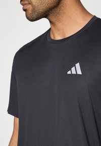 Camiseta deportiva negra con cuello redondo, mangas cortas y un logotipo de la marca reflectante en la esquina superior izquierda. Textura suave.