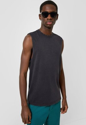 Mann trägt schwarzes ärmelloses Shirt, türkisfarbene Hose, schwarze Sonnenbrille und kleine Creolen, steht vor einem hellgrauen Hintergrund.