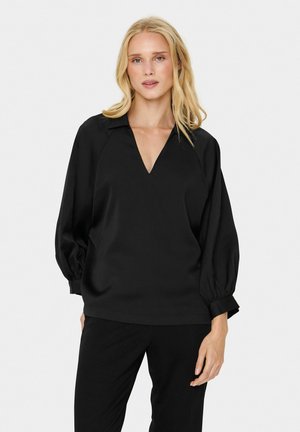 Black blouse with a V-neck, raglan sleeves, and gathered cuffs. Smooth texture, relaxed fit, and minimalistic design. Worn with black pants.