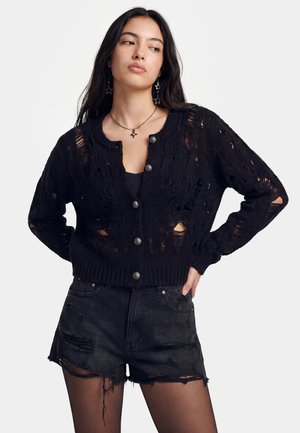 Ung kvinde iført sort slidt cardigan med knapper, sort top, revnede sorte denimshorts, stjerneformede øreringe og halskæde, stående med hænderne på hofterne.