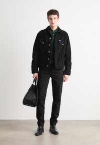 Ung mand iført sorte jeans, sort jakke, mørkegrøn skjorte, sorte støvler, der holder en sort taske, stående mod en ensartet lys væg.