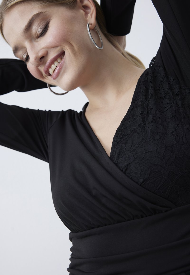 Top avvolgente nero con un pannello in pizzo testurizzato su un lato, realizzato in un tessuto morbido con vita arricciata e maniche lunghe, progettato per un look aderente.