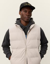 Επενδεδυμένο, αμάνικο γιλέκο σε ανοιχτό μπεζ, με μπροστινό φερμουάρ και διακριτικό λογότυπο. Φοριέται πάνω από ένα σκούρο φούτερ με κορδόνι.