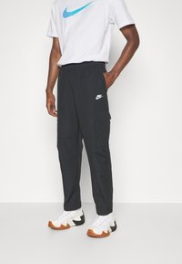 Person klädd i svarta Nike cargo-byxor, vit t-shirt med blå logotyp och vita sneakers med bruna och svarta sulor som står mot en vit bakgrund.