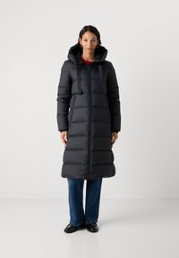 Zwarte pufferjack met een capuchon, voorzien van een rits aan de voorkant, linten en zijzakken; knie-lengte met een gewatteerde textuur. Gepaard met blauwe jeans.