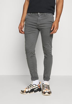 Mann trägt schlanke graue Hose, weiße Socken und weiße, schwarze und orangefarbene Sportschuhe und steht vor einem weißen Hintergrund.