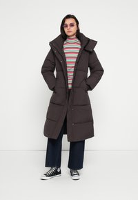 Dugi, tamnocrni smeđi jakni za snijeg s visokim ovratnikom, patentnim zatvaračem i kopčama na pritisak. Nosi se preko višebojnog prugastog rolka i širokih traperica.