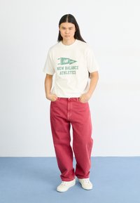 Camiseta crema con gráfico verde de "New Balance Athletics", combinada con pantalones rojos de corte amplio y zapatillas blancas sobre un suelo azul.