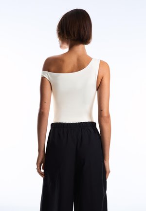 Vrouw met kort bruin haar die een witte off-shoulder top en losse zwarte broek draagt, staand met haar rug naar een witte achtergrond.