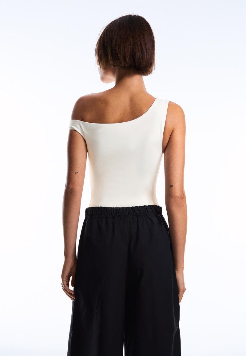 Femme aux cheveux courts marron, portant un haut blanc épaules dénudées et un pantalon noir ample, debout de dos sur un fond blanc.