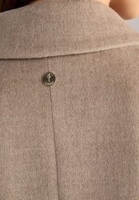 Béžový vlněný kabát s překládaným límcem a kulatým zlatým knoflíkem se znakem jelena na zadní straně, vykazující jemnou texturu a čisté linie.