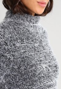 Maglione grigio e bianco fluffy con colletto alto. Tessuto testurizzato, morbido con un aspetto peloso. Vista ravvicinata del collo e del motivo.