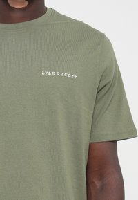 T-shirt en coton vert olive avec des manches courtes et un col ras du cou. Présente un logo "LYLE & SCOTT" brodé en blanc sur la poitrine gauche.