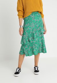 Woman wearing a mustard yellow sweater, green floral midi skirt with ruffles, silver cuff bracelet, and black and white sneakers.