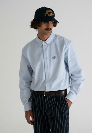 Light blue button-up shirt with a small navy embroidered logo, paired with striped navy pants and a black cap featuring golden text.